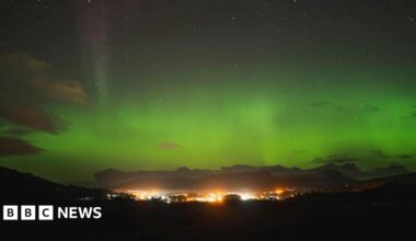 The aurora, also known as the Northern Lights, glows green above a brightly lit community in the Isle of Skye.