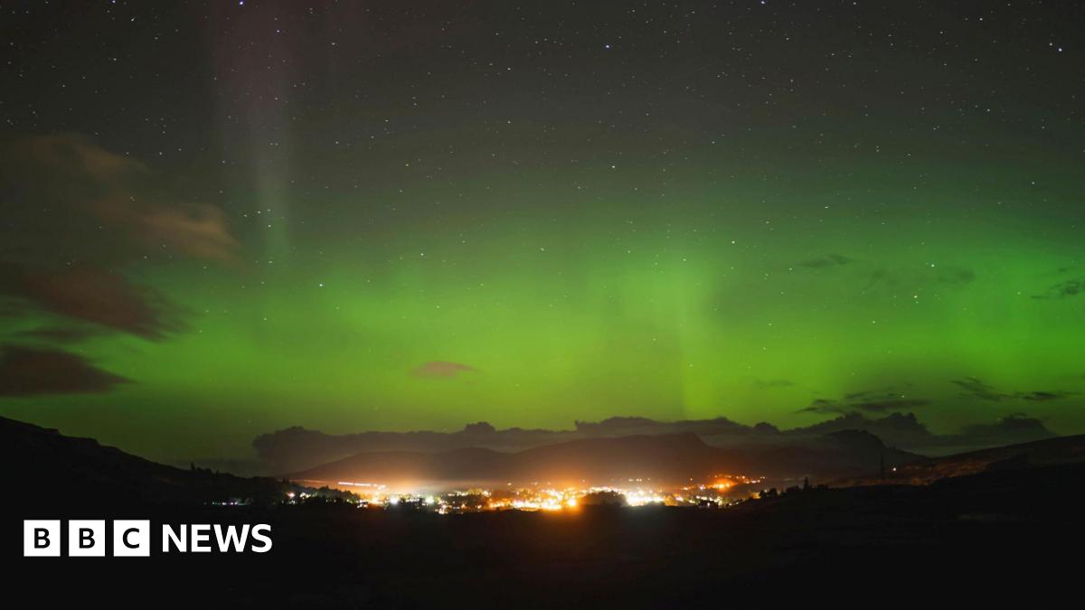 The aurora, also known as the Northern Lights, glows green above a brightly lit community in the Isle of Skye.