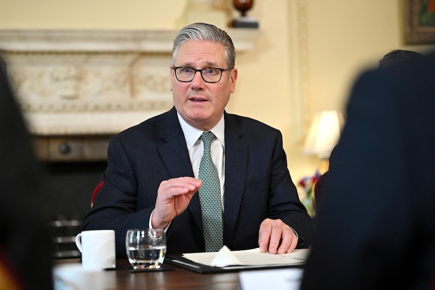 Keir Starmer wearing a black suit and teal tie, mid-speech while sitting at a wooden table and gesturing with his right hand.