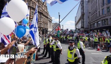 Police officers in high-visibility vests stand between two groups of protesters on a city street. To the left, people hold Israeli flags, a Union Jack, and white and blue balloons. To the right, behind a metal barrier, a separate crowd holds Palestinian flags and various protest placards. Historic buildings and a bright blue sky are visible in the background.