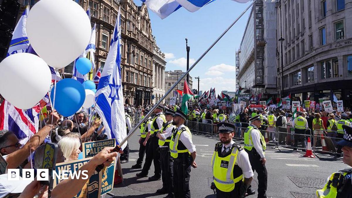 Police officers in high-visibility vests stand between two groups of protesters on a city street. To the left, people hold Israeli flags, a Union Jack, and white and blue balloons. To the right, behind a metal barrier, a separate crowd holds Palestinian flags and various protest placards. Historic buildings and a bright blue sky are visible in the background.