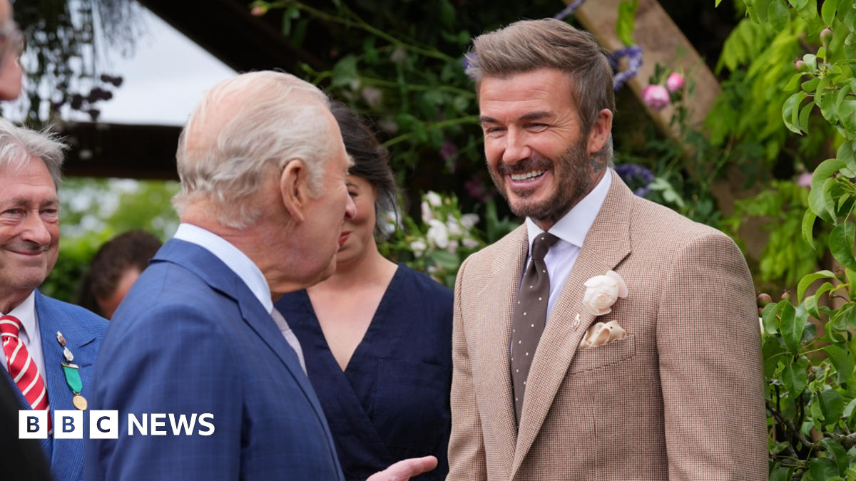 A main with short blonde hair and a light brown suit jacket and brown tie standing in front of a green hedge and talking to a man with short grey hair and a blue jacket