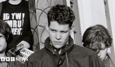 The black and white image shows a protest taking place on the steps of the then Welsh Office. On the left a campaigner is speaking into a loud-speaker. On the right a number of young people are listening on. They include Rhun ap Iorwerth who is holding a placard.