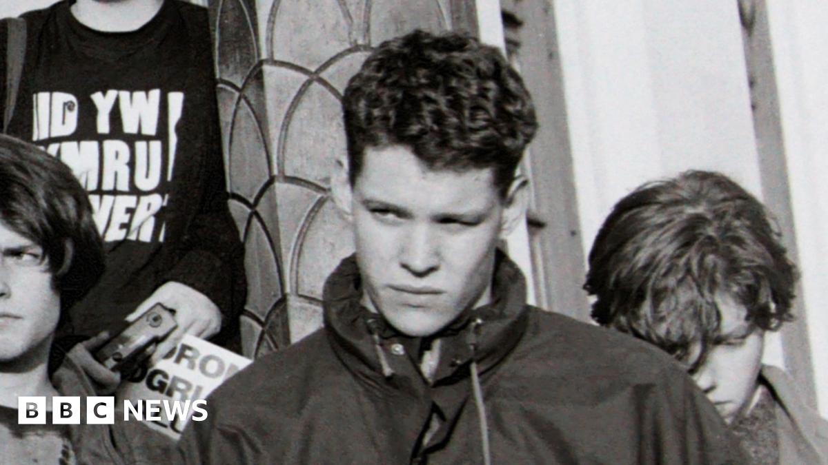 The black and white image shows a protest taking place on the steps of the then Welsh Office. On the left a campaigner is speaking into a loud-speaker. On the right a number of young people are listening on. They include Rhun ap Iorwerth who is holding a placard.