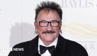 Paul Chuckle at a red carpet event. It is a close crop of his face. He has a brown moustache, brown hair sticking up around his head and black square glasses. He is smiling and wears a white shirt, black bowtie and black jacket.