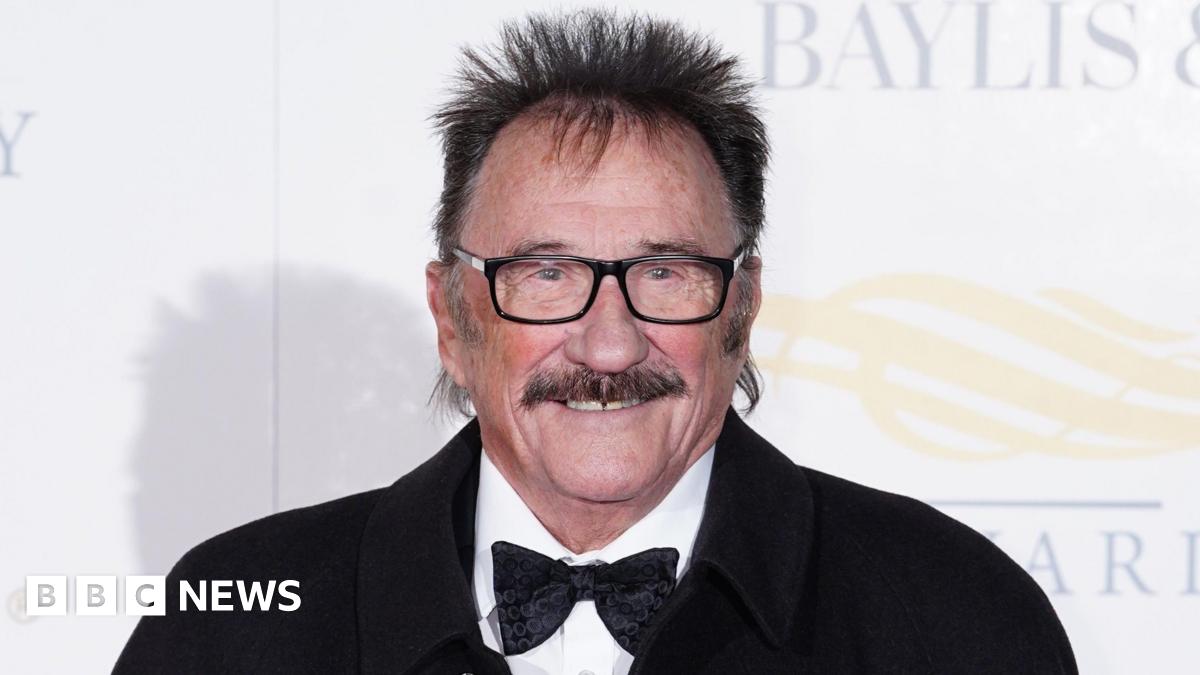 Paul Chuckle at a red carpet event. It is a close crop of his face. He has a brown moustache, brown hair sticking up around his head and black square glasses. He is smiling and wears a white shirt, black bowtie and black jacket.
