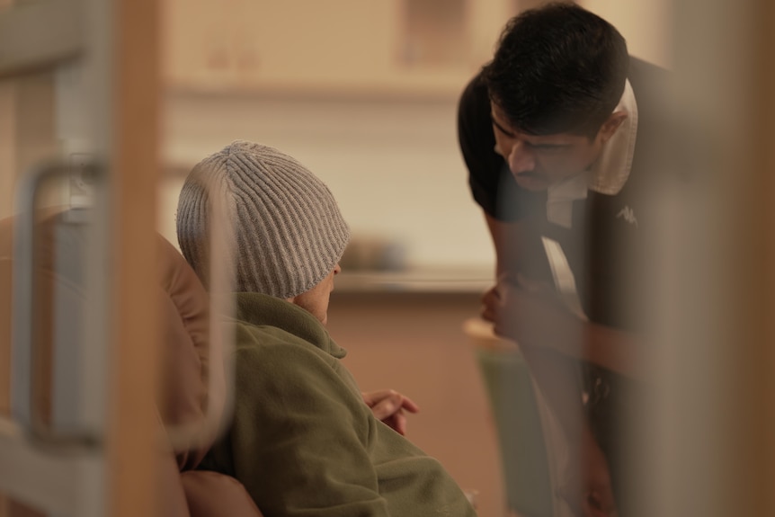 An aged care staffer speaks to a resident sitting on a recliner.