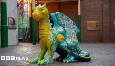 A large dragon sculpture that is green, yellow and blue, with yellow claws. It has drawn stickers of people on it, with a yellow circular logo that reads "Compton Care"