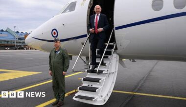John Healey in a white shirt, black suit and red tie, seen exiting an RAF plane at an airport as a soldier stands to attention nearby