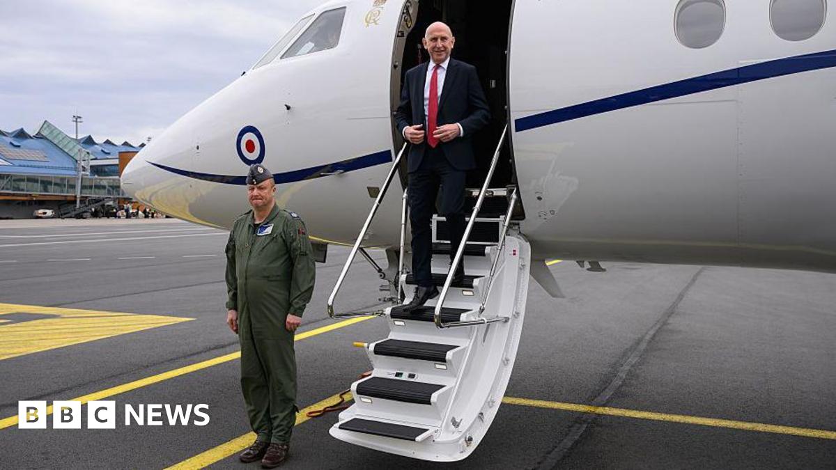John Healey in a white shirt, black suit and red tie, seen exiting an RAF plane at an airport as a soldier stands to attention nearby