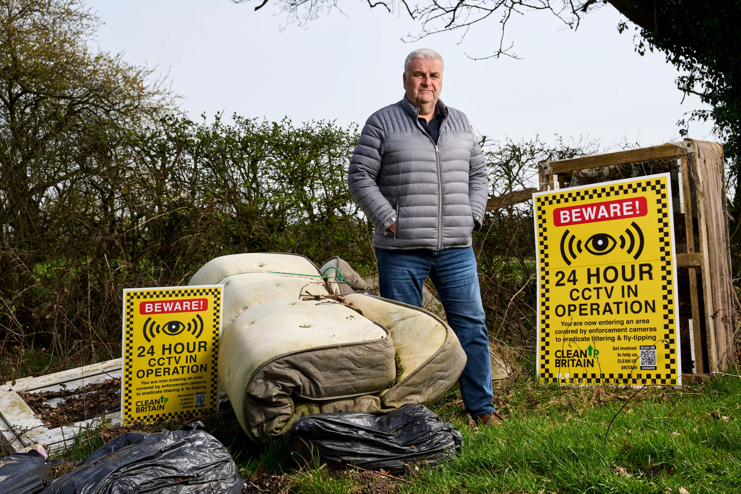 John Read, Co-Founder of Clean Up Britain, stands near fly-tipped household waste with "24 Hour CCTV in Operation" signs.