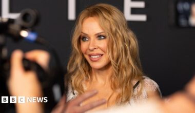 A woman (Kylie Minogue) with wavy blonde hair smiles while being photographed at an event, with blurred cameras and people in the foreground.