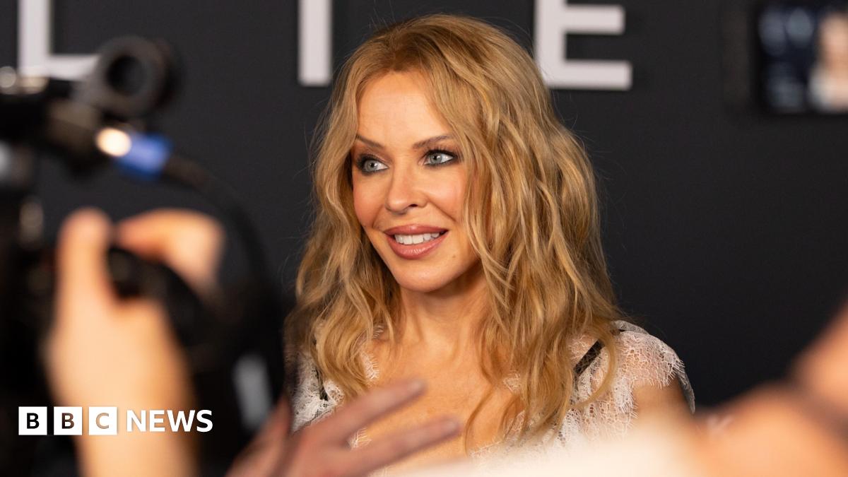 A woman (Kylie Minogue) with wavy blonde hair smiles while being photographed at an event, with blurred cameras and people in the foreground.