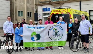 Two cyclists standing for a photo outside a building with people cheering them on stood between them. An ambulance is behind them.