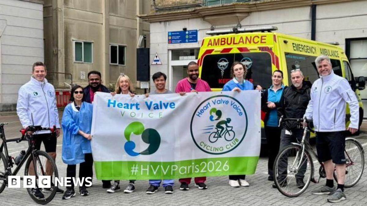 Two cyclists standing for a photo outside a building with people cheering them on stood between them. An ambulance is behind them.