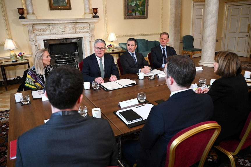 Keir Starmer in a black suit and blue tie sitting at a wooden table with men and women in black suits.