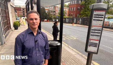 A man in a blue long sleeved-top standing by a bus stop on Golders Green Road.