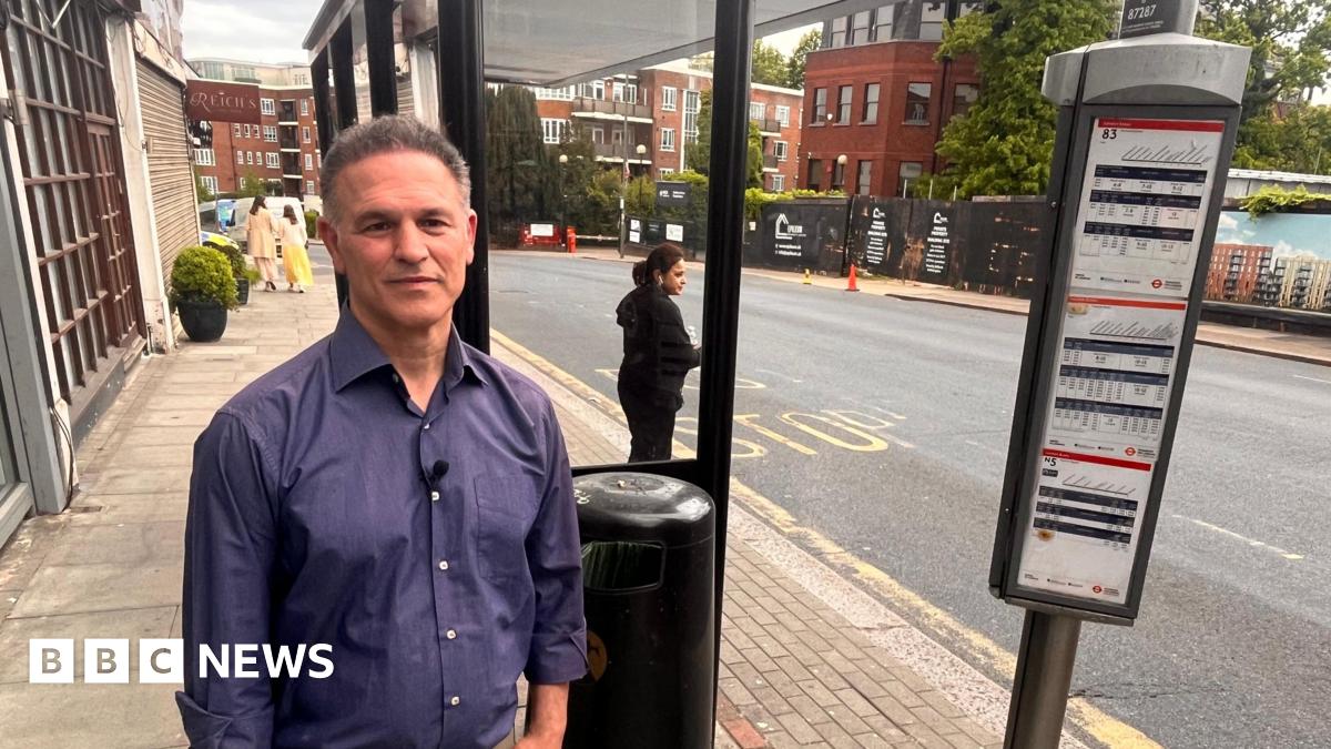 A man in a blue long sleeved-top standing by a bus stop on Golders Green Road.