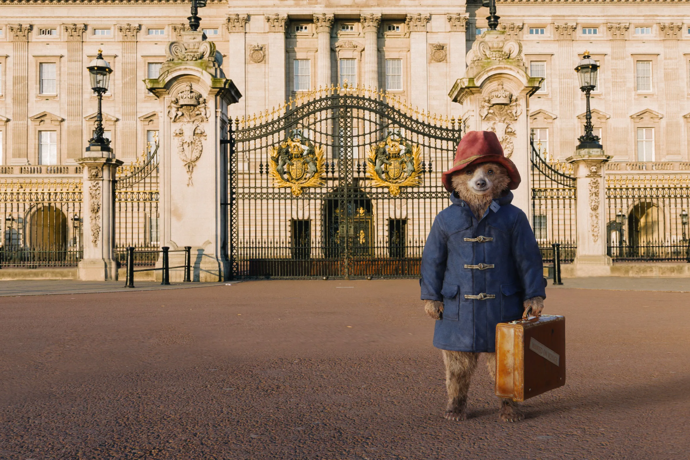 Paddington Bear stands with a suitcase in front of Buckingham Palace.