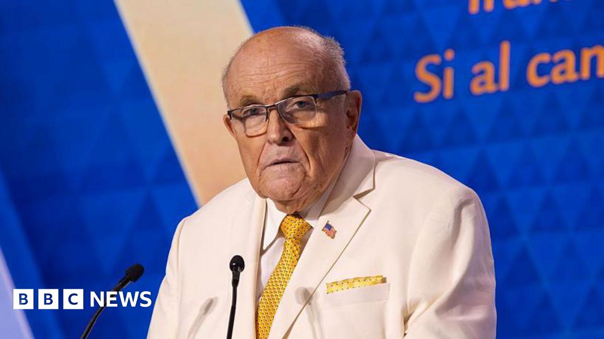 Giuliani in center with blue suit jacket over white medical brace, crosses his hands and looks down. He is flanked by Kash Patel and Howard Lutnick