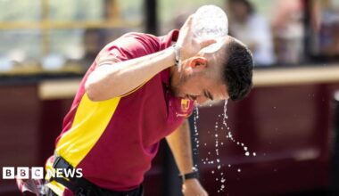 A man with short dark hair and a short beard pours water from a bottle over his neck. He is wearing a red and yellow top and has a belt around his hip. The background behind him is blurred but appears to be a bus.