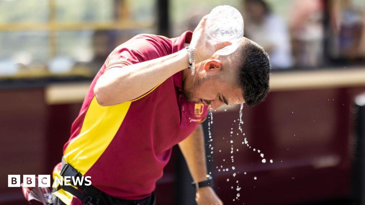 A man with short dark hair and a short beard pours water from a bottle over his neck. He is wearing a red and yellow top and has a belt around his hip. The background behind him is blurred but appears to be a bus.