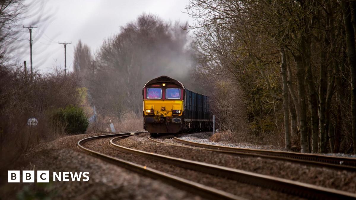 A cargo train approaching in the distance on a track. It has a yellow cab with a red top. There is mist coming off the roof. Trees line the track.