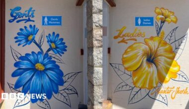 Close-up of two painted walls by public toilets side by side. The left door is labelled “Gents Hommes” and decorated with large blue flowers; the right door is labelled “Ladies Dames” and decorated with large yellow flowers. The artwork is painted directly onto the wall.