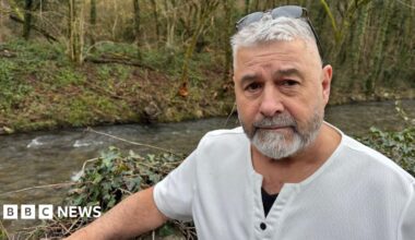 A man look back at the camera as he gestures with one hand towards a pile of boxes, a child's green dinosaur chair, a seed spreader and plastic bags for life. He has short white hair and a clipped salt and pepper beard. he is wearing a linen elbow length shirt open at the neck