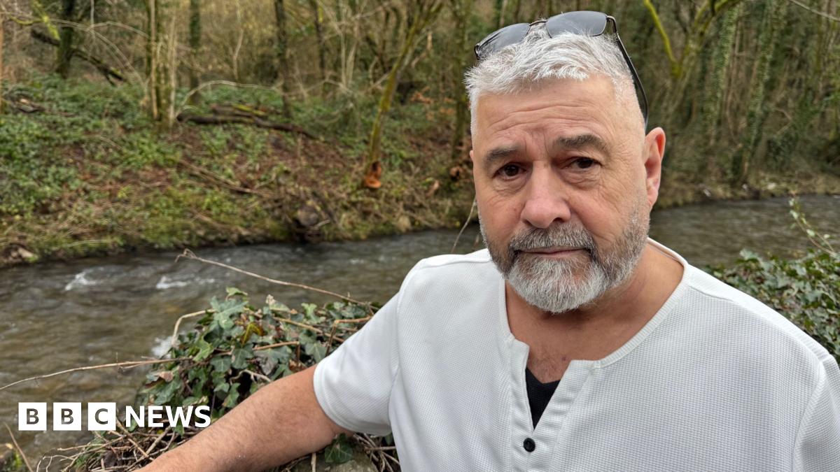 A man look back at the camera as he gestures with one hand towards a pile of boxes, a child's green dinosaur chair, a seed spreader and plastic bags for life. He has short white hair and a clipped salt and pepper beard. he is wearing a linen elbow length shirt open at the neck