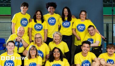 A group of 13 people smiling and posing together indoors, wearing bright yellow T-shirts with a blue circular logo that reads "JOY MAKER". They’re arranged in two rows, some seated and others standing, against a grey backdrop in what appears to be a gym or community centre.