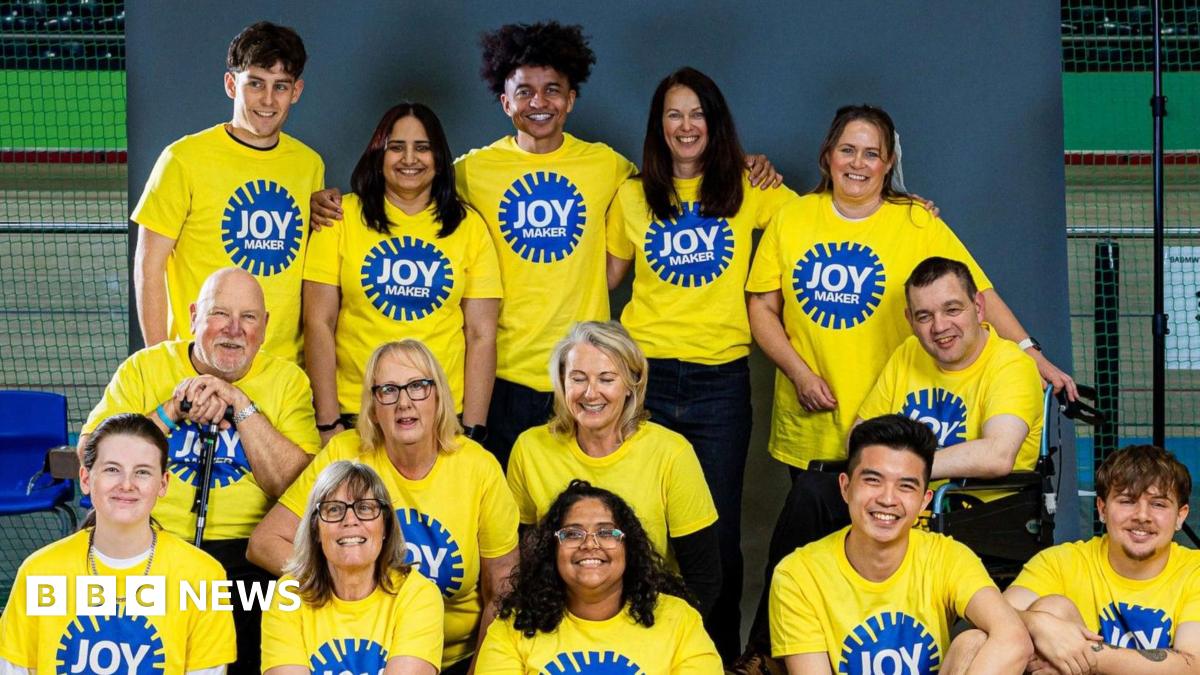 A group of 13 people smiling and posing together indoors, wearing bright yellow T-shirts with a blue circular logo that reads "JOY MAKER". They’re arranged in two rows, some seated and others standing, against a grey backdrop in what appears to be a gym or community centre.