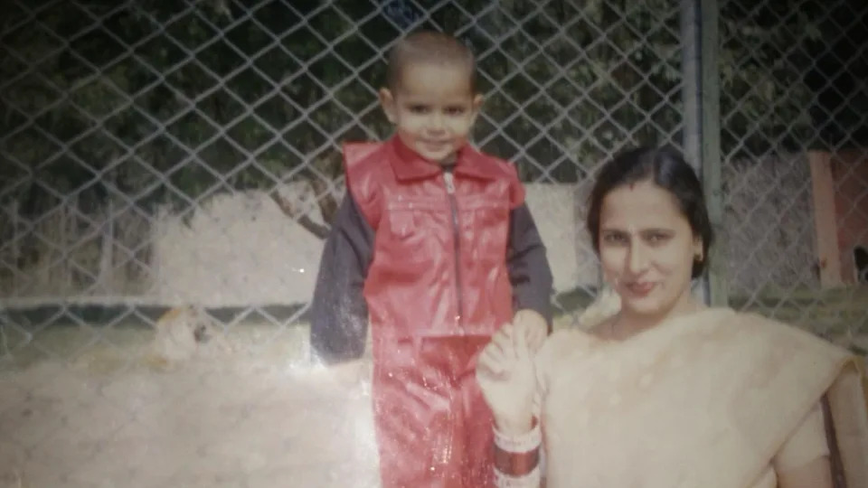 A slightly faded photograph shows a small child (Tushar Kumar) in a bright red outfit standing beside an woman near a chain-link fence. The adult wears a light-coloured outfit and jewellery, while the child’s shiny red clothing stands out against the muted background. Behind them, a light-coloured animal can be seen inside the enclosure.
