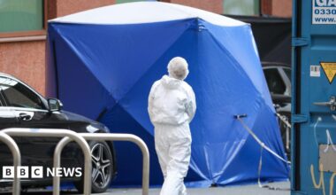 A blue forensic tent is erected in the street with an officer dressed in white overalls walking towards to. A black car is parked next to the tent