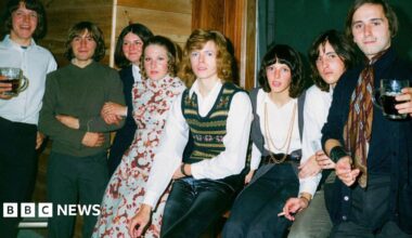 David Bowie, who has curly dark blonde hair, poses for a photo backstage at a club surrounded by seven teenagers including four boys and three girls.