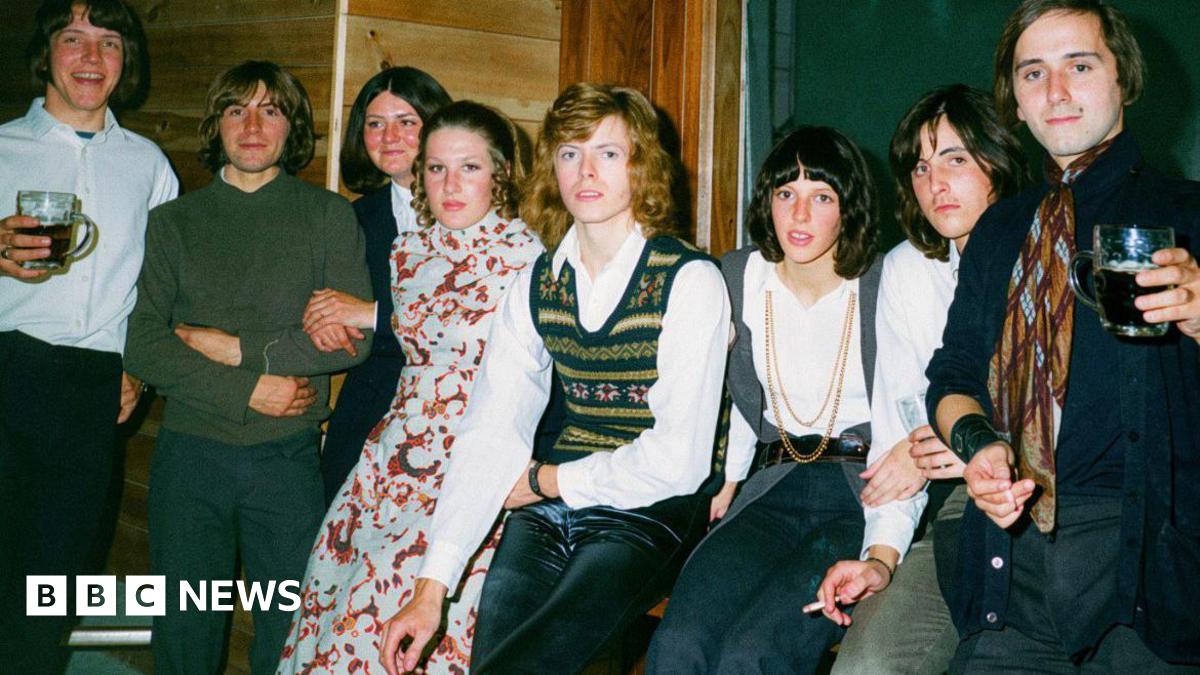 David Bowie, who has curly dark blonde hair, poses for a photo backstage at a club surrounded by seven teenagers including four boys and three girls.