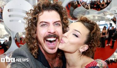 A man with curly brown hair, smiling widely at the camera, while a woman kisses him on the cheek