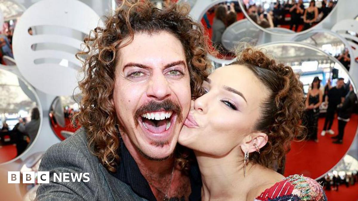 A man with curly brown hair, smiling widely at the camera, while a woman kisses him on the cheek
