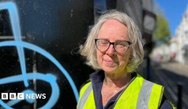 Emma Humphreys standing in Trafalgar Street, Brighton, next to a wall with blue spray paint on it. She has grey hair and is wearing a high vis vest
