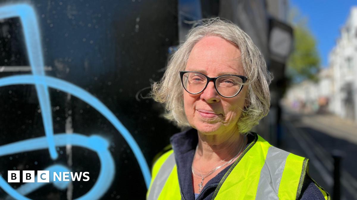 Emma Humphreys standing in Trafalgar Street, Brighton, next to a wall with blue spray paint on it. She has grey hair and is wearing a high vis vest