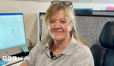 Angela is sat in an office on a black leather chair. She is wearing a grey polo shirt with the West Wales Electrical Solutions logo which is a yellow light bulb. She has her arm rested on a black desk. On the desk is a white desk top computer and a white keyboard.