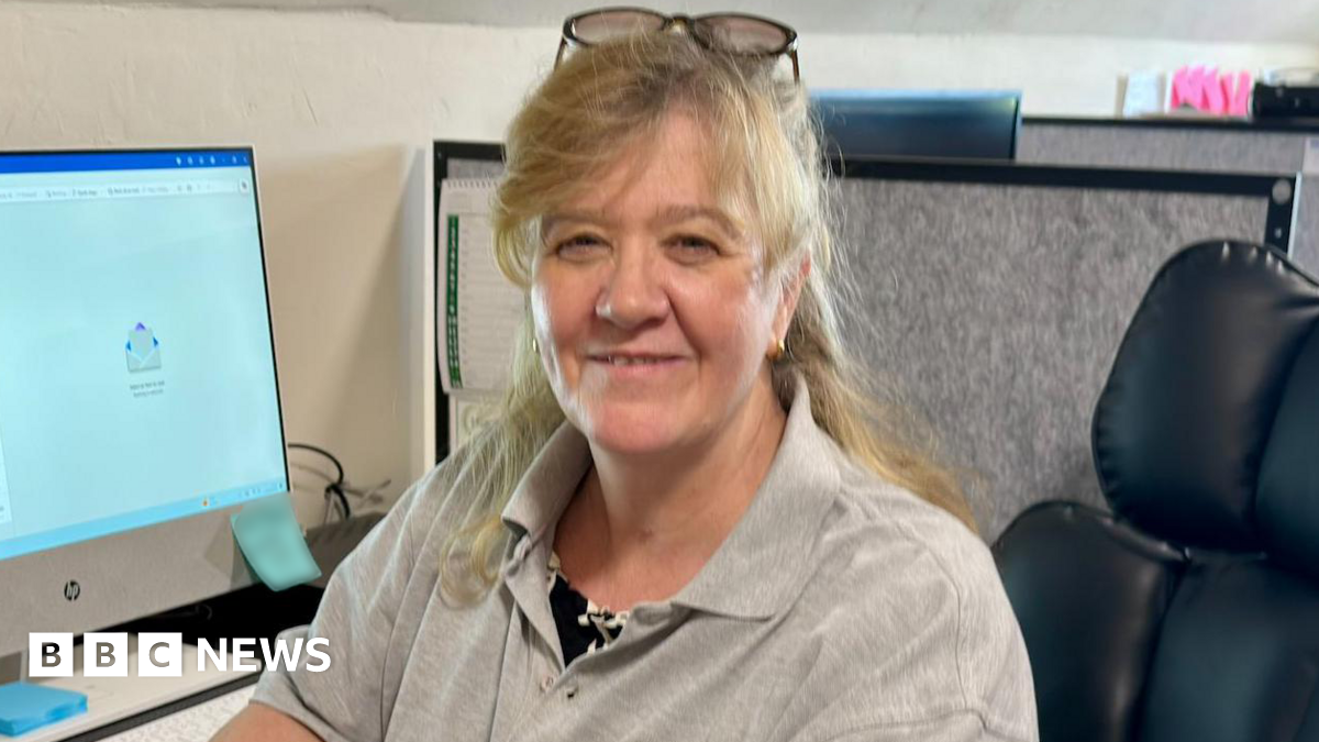 Angela is sat in an office on a black leather chair. She is wearing a grey polo shirt with the West Wales Electrical Solutions logo which is a yellow light bulb. She has her arm rested on a black desk. On the desk is a white desk top computer and a white keyboard.