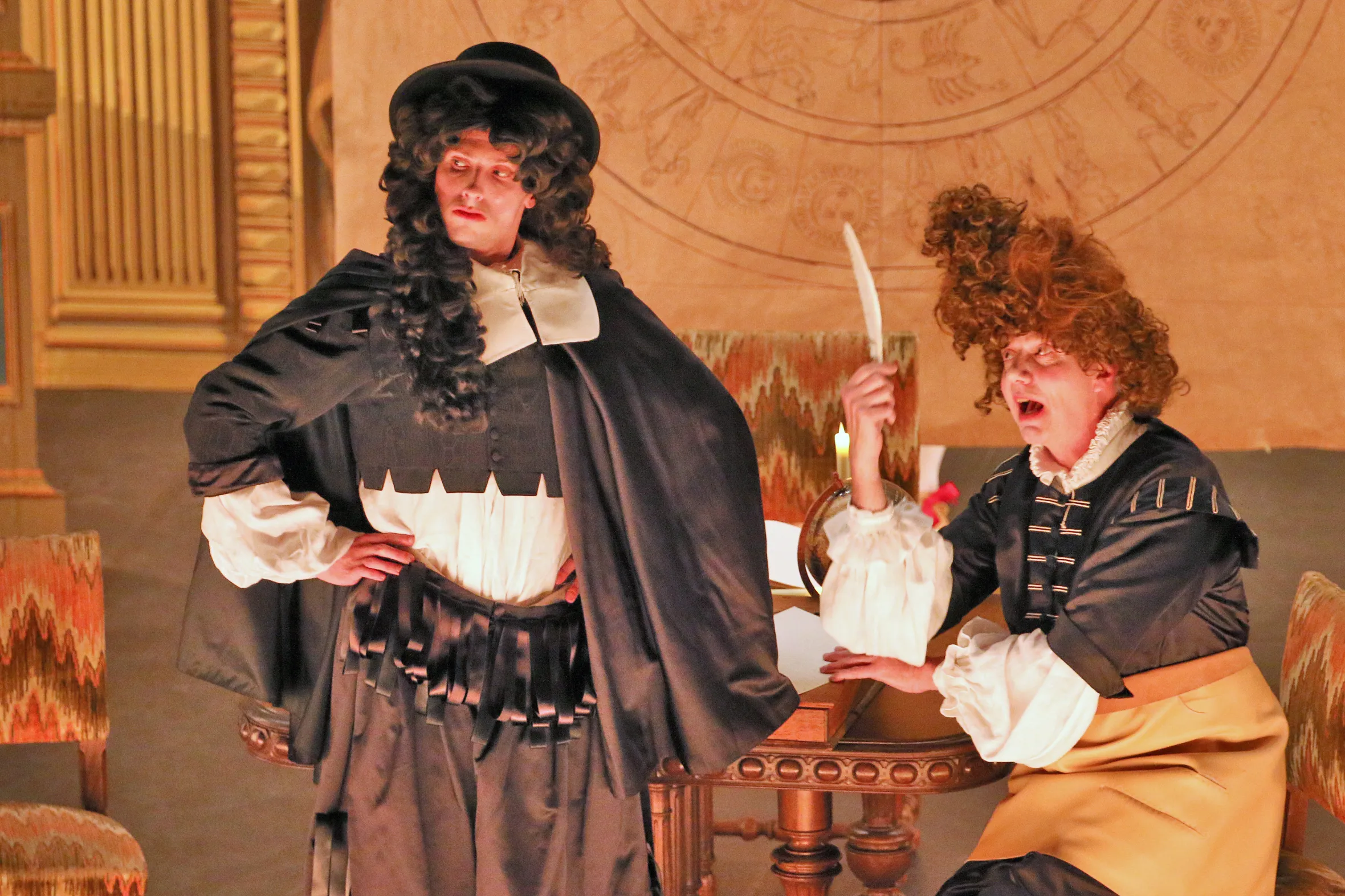 Two actors in period costumes from Molière's play, "The Astrologer or False Omens," one standing with hands on hips, the other seated at a table with a quill.