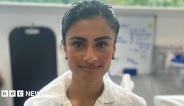 Close-up of Taibar Akhtar standing in the hospital ward in a white uniform with a plastic apron over the top. She is smiling at the camera and has dark hair tied back.
