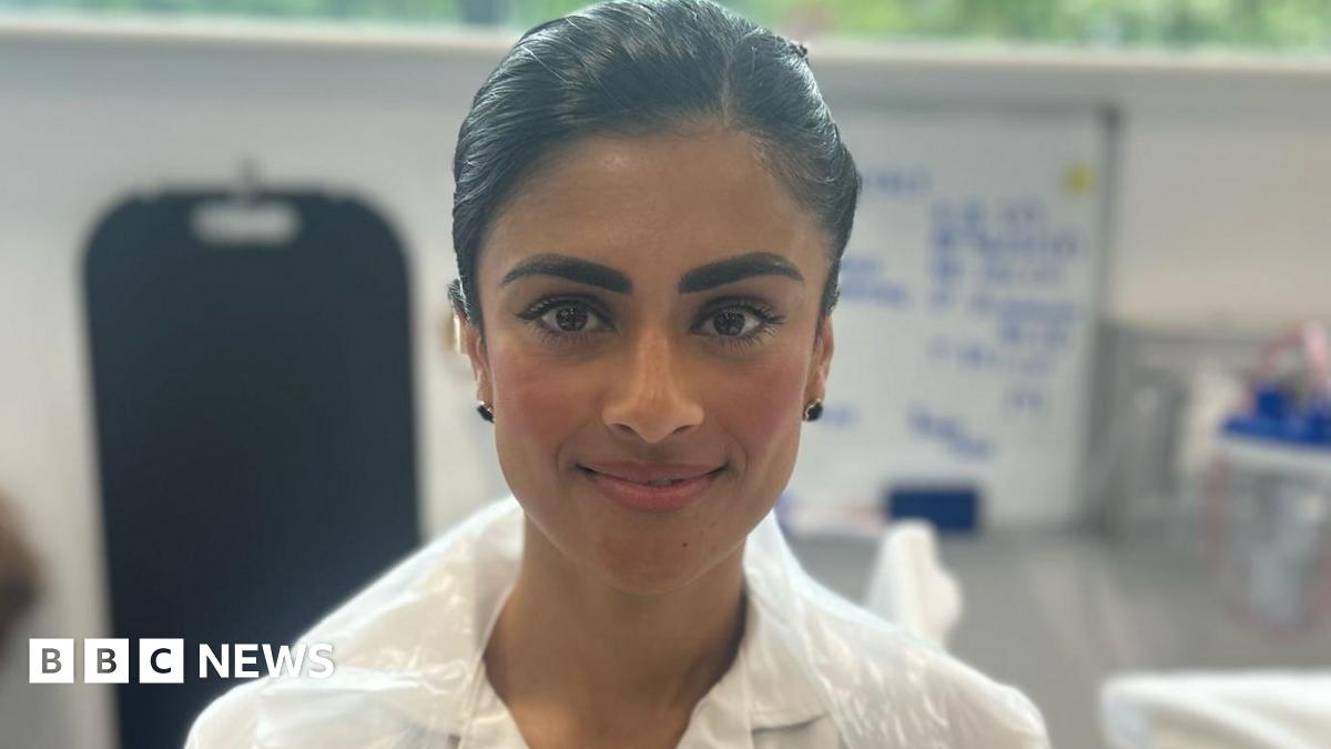 Close-up of Taibar Akhtar standing in the hospital ward in a white uniform with a plastic apron over the top. She is smiling at the camera and has dark hair tied back.