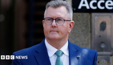 A man with short, cropped grey hair looks to the left of the camera. He has black-rimmed glasses and is wearing a blue suit, white shirt and green patterned tied. He is standing outside a building, a gateway can be seen, out of focus, behind him