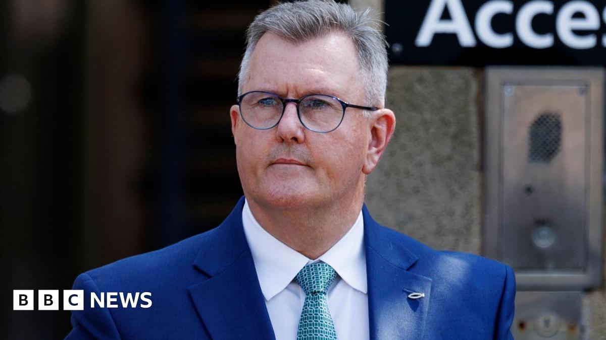 A man with short, cropped grey hair looks to the left of the camera. He has black-rimmed glasses and is wearing a blue suit, white shirt and green patterned tied. He is standing outside a building, a gateway can be seen, out of focus, behind him