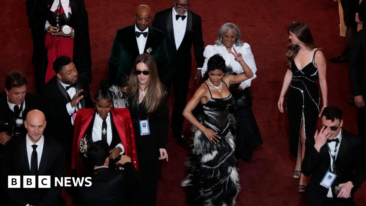 Actor and singer Teyana Taylor wearing a fitted gown of black and white feathers as she shimmies down the red carpet of the Acadamy Awards surrounded by other attendees.