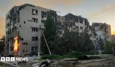 A large grey boxy building is visibly damaged and surrounded by debris and rubble. Flames are visible on one side of the building.
