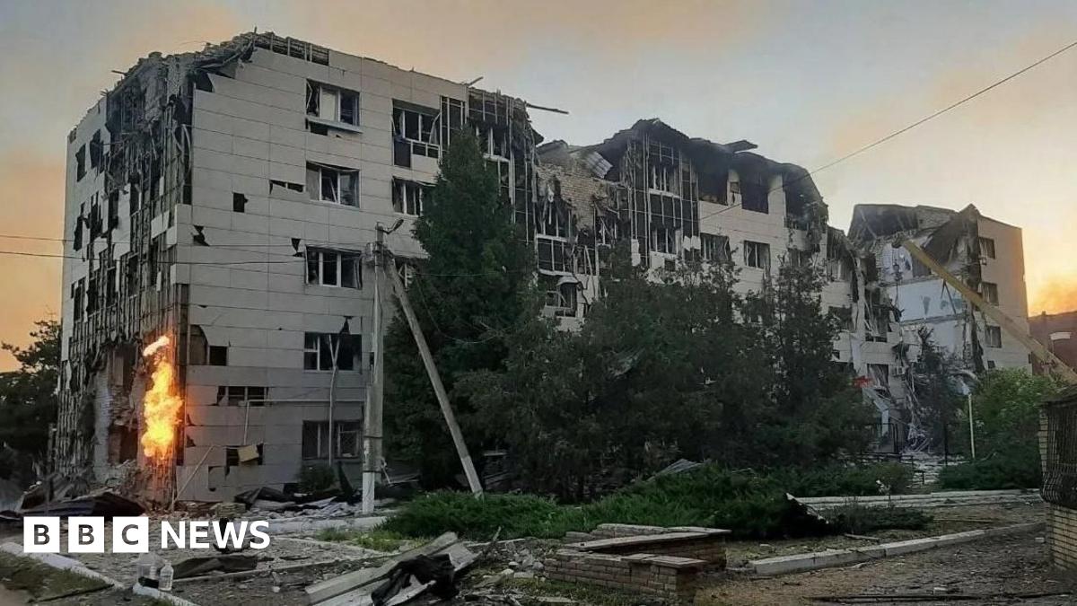A large grey boxy building is visibly damaged and surrounded by debris and rubble. Flames are visible on one side of the building.